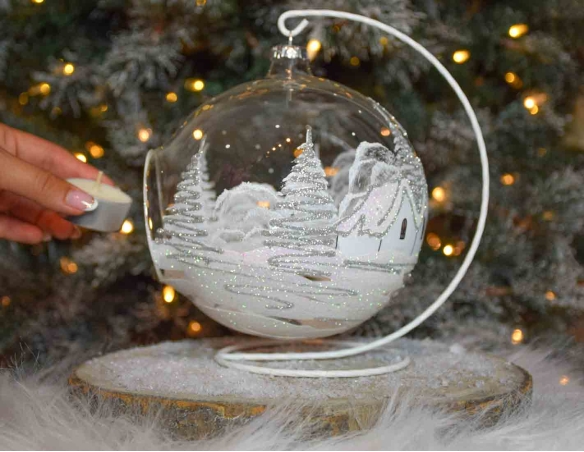 Grande boule bougeoir de Noël en verre soufflé avec un décor village blanc et paillettes argentées. Diamètre 15cm.